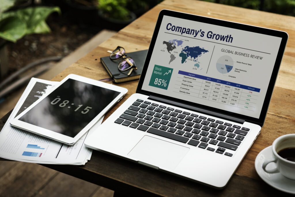 Laptop displaying a report titled "Company's Growth" with global business statistics, alongside a tablet showing a clock, papers with charts, and glasses, symbolizing IT support and business strategy evolution.